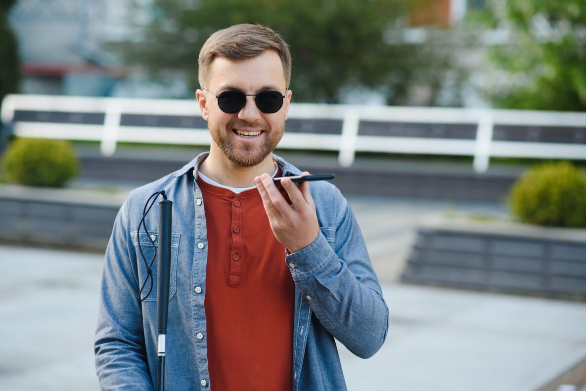 A man with a cane and dark glasses using a smartphone.