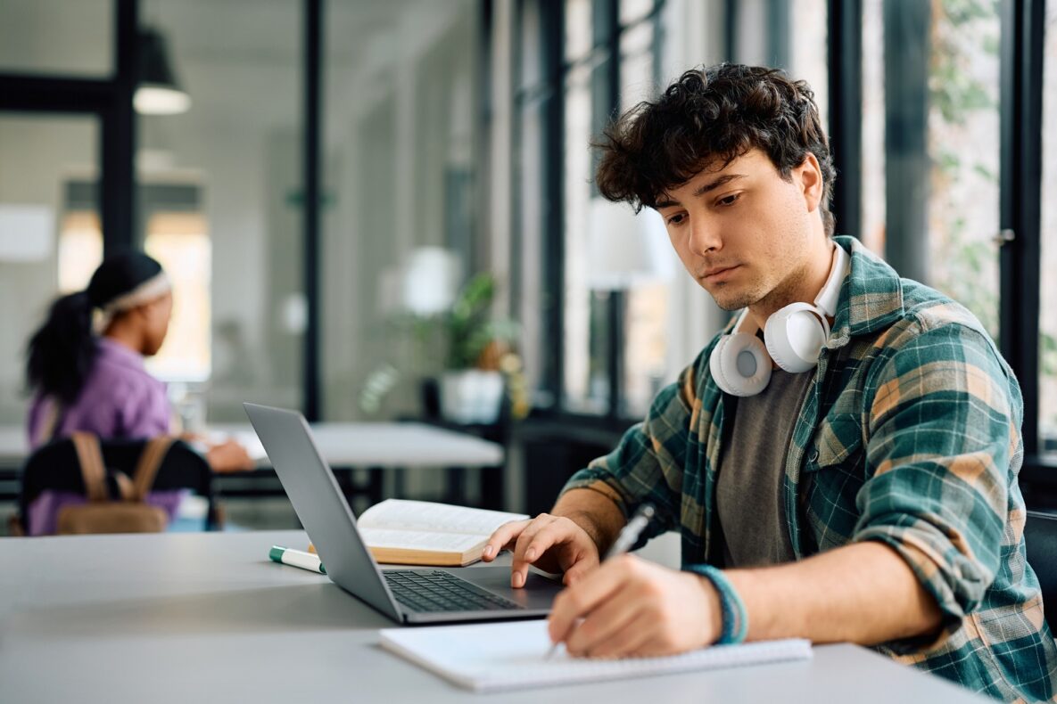 University student writing while using laptop and studying in the classroom.
