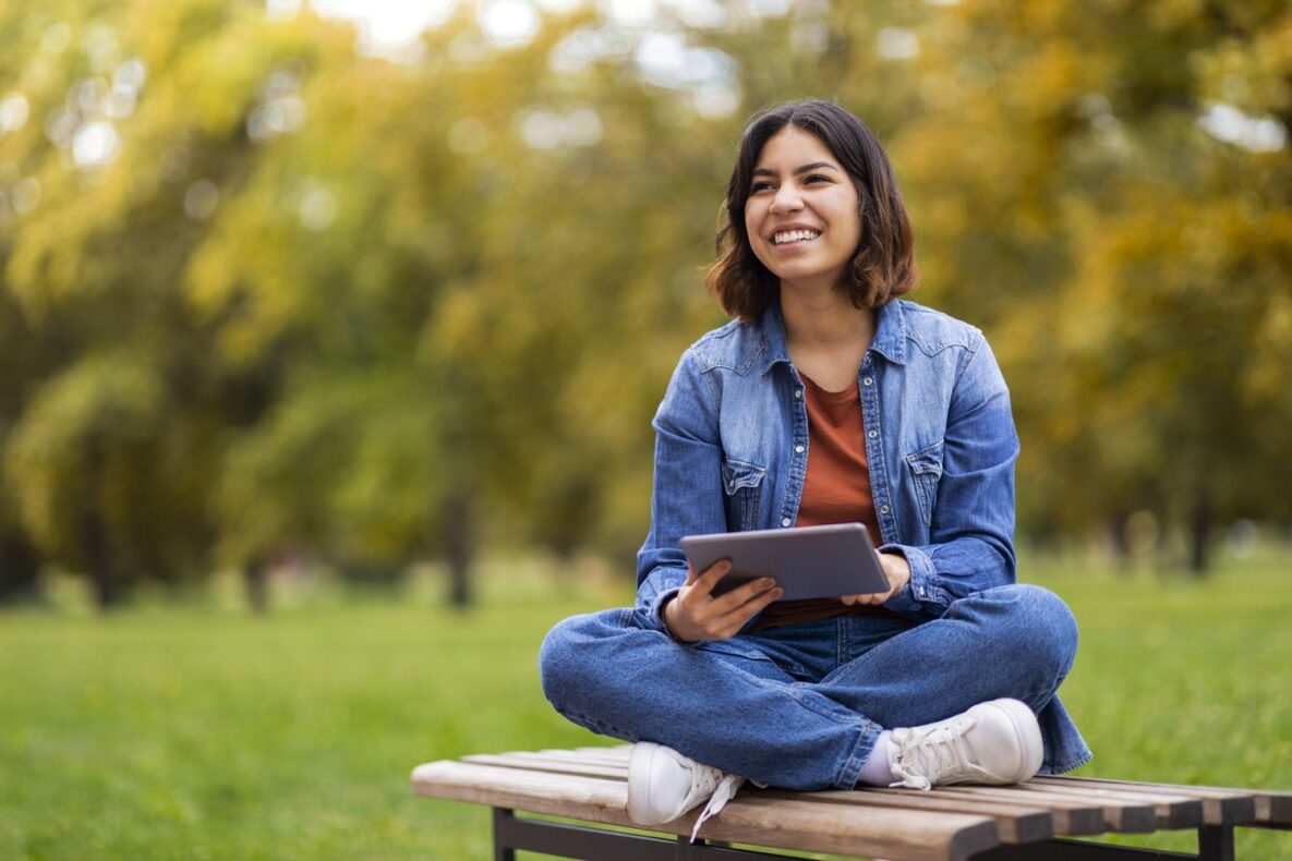 Jeune femme arabe heureuse et belle se relaxant avec une tablette numérique en plein air, femme du Moyen-Orient joyeuse assise sur un banc dans un parc et utilisant un gadget moderne, naviguant sur Internet ou consultant les réseaux sociaux